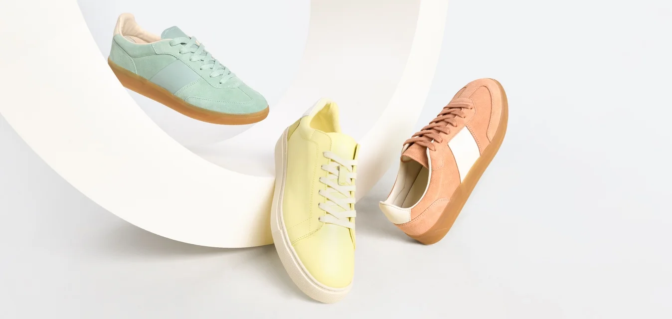Tre sneaker casual nei colori pastello menta, giallo e pesca disposte attorno a una struttura astratta bianca e curvata su uno sfondo chiaro.