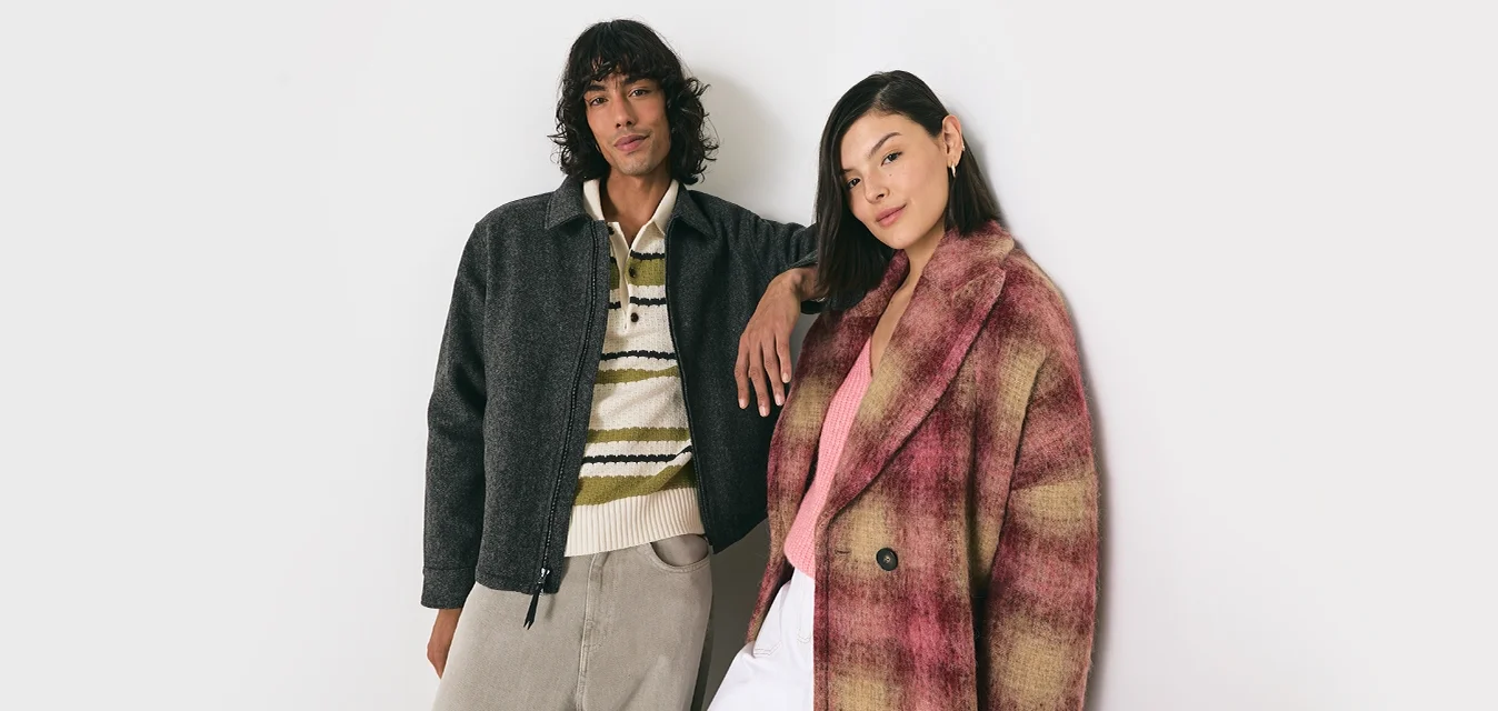 Due giovani adulti in piedi contro un muro bianco, uno indossa una giacca scura e un maglione a righe, l'altro un cappotto a quadri rosa e beige.