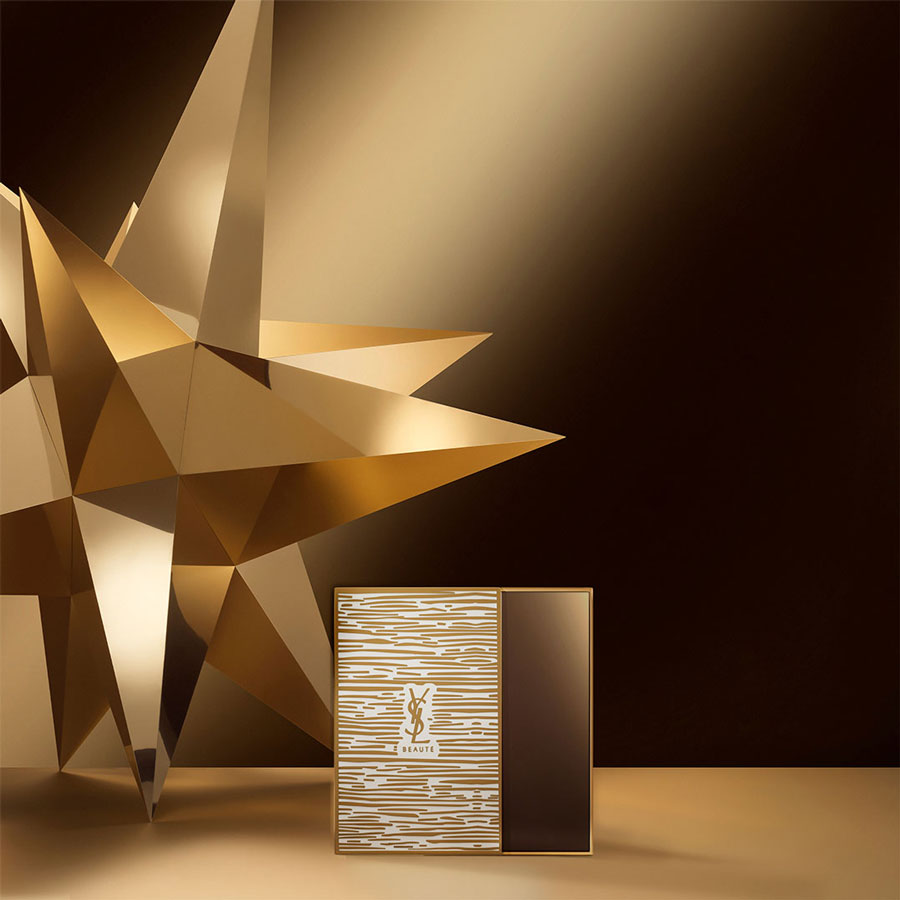 Uno specchio dorato YSL Beauté, con eleganti motivi a strisce bianche e il logo YSL, è esposto accanto a una prominente stella dorata astratta su uno sfondo scuro e sfumato.