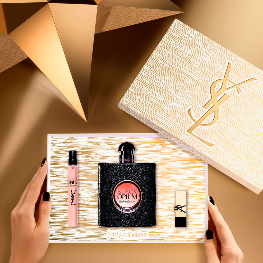 Un paio di mani con smalto nero tengono una confezione regalo Yves Saint Laurent aperta contenente un profumo Black Opium a grandezza naturale, un roll-on Black Opium mini e un rossetto nero e oro, con un'altra grande scatola YSL sullo sfondo, il tutto su un'ambientazione geometrica dorata.