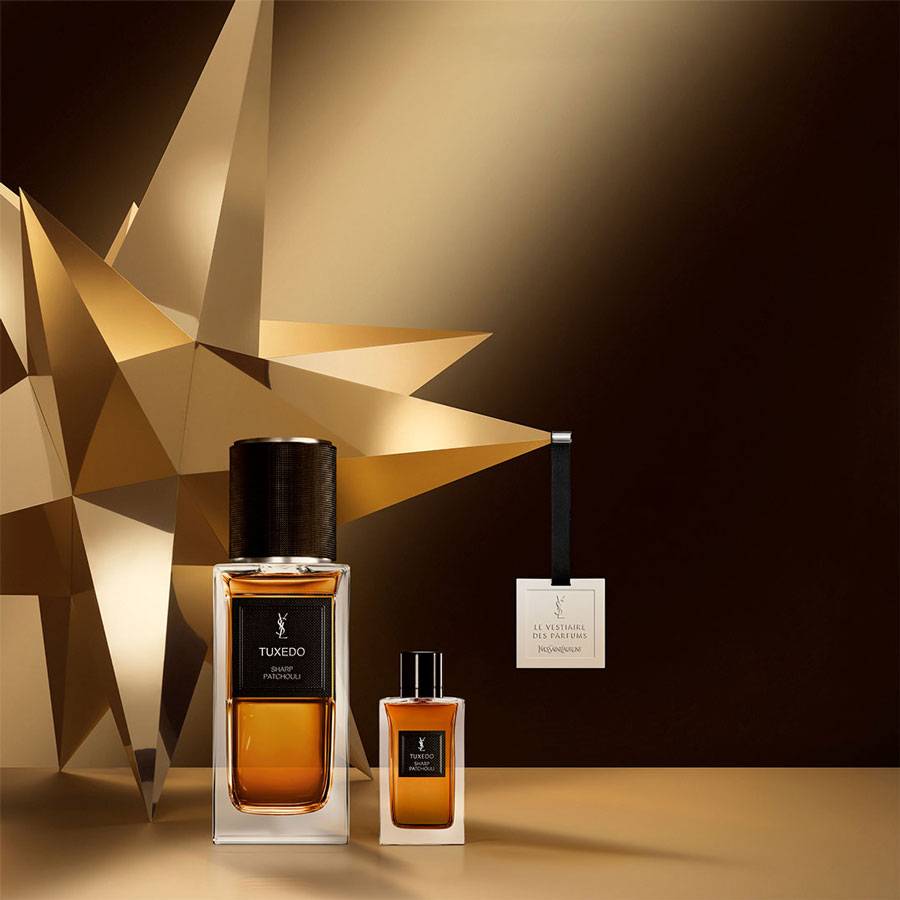 Due flaconi del profumo YSL Le Vestiaire Des Parfums "Tuxedo Sharp Patchouli", insieme a un'etichetta profumata appesa, sono esposti su una superficie dorata con decorazioni geometriche a stella sullo sfondo.