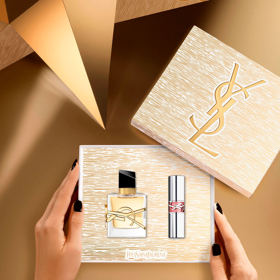 Un paio di mani con smalto nero tengono una confezione regalo Yves Saint Laurent aperta contenente una bottiglia di profumo Libre e un balsamo labbra rosa, con un'altra scatola YSL più grande sullo sfondo, il tutto su un'ambientazione geometrica dorata.