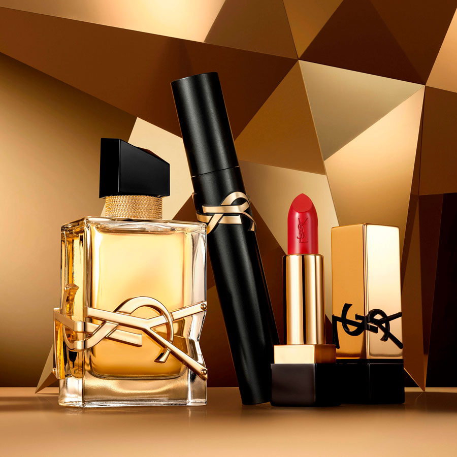 Una composizione del profumo Yves Saint Laurent Libre, un mascara nero e un rossetto rosso, tutti con accenti dorati, disposti su un lussuoso sfondo geometrico dorato.