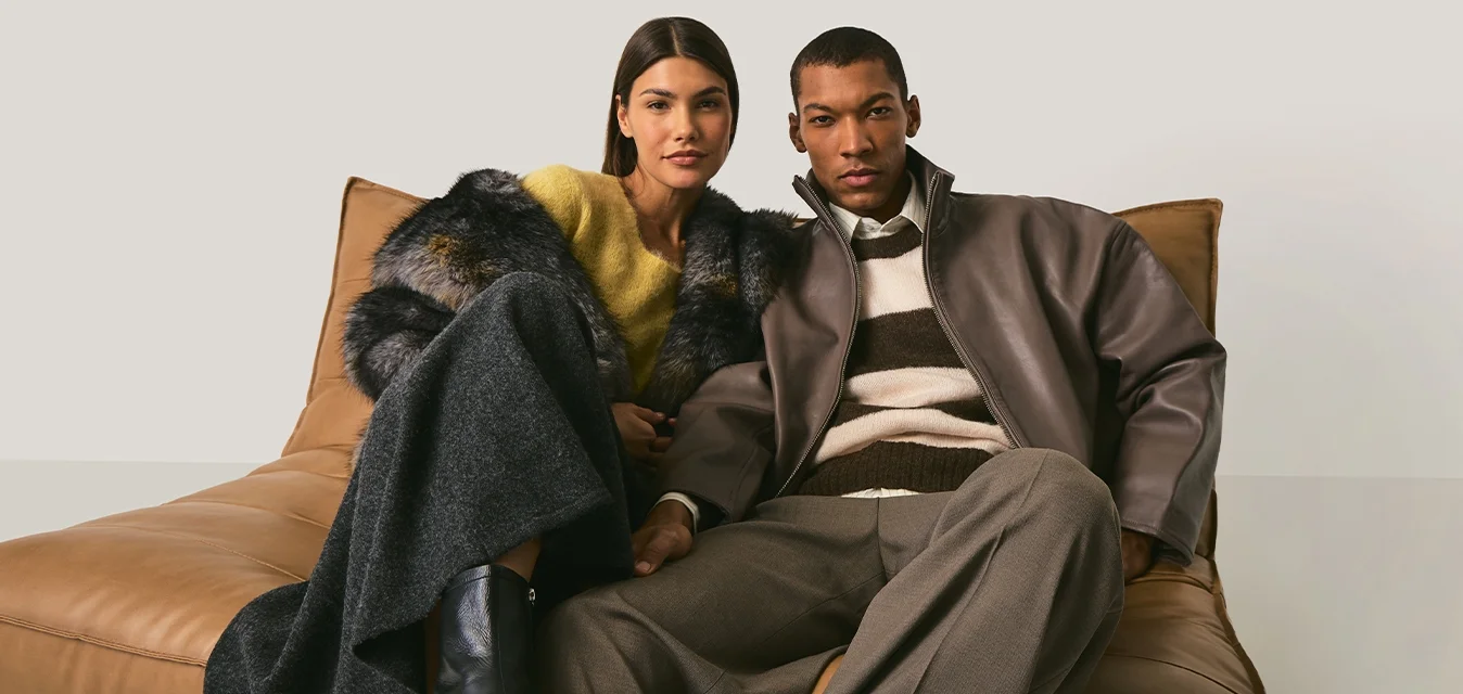 Due persone sedute vicine su un divano in pelle color cuoio, vestite con outfit autunnali alla moda con cappotti e stivali, su uno sfondo beige semplice.