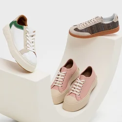 Sneaker casual bianchi con accenti verdi, rosa chiaro e texture intrecciate marroni, esposte su una piattaforma curva bianca.