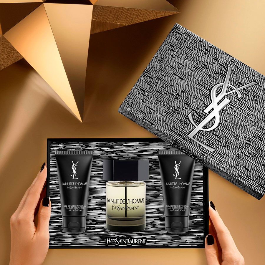 Un paio di mani con smalto nero tengono una confezione regalo Yves Saint Laurent aperta contenente una bottiglia di profumo La Nuit de L'Homme e due tubi di gel doccia abbinato, con una grande scatola YSL in bianco e nero sullo sfondo, il tutto su un'ambientazione geometrica dorata.