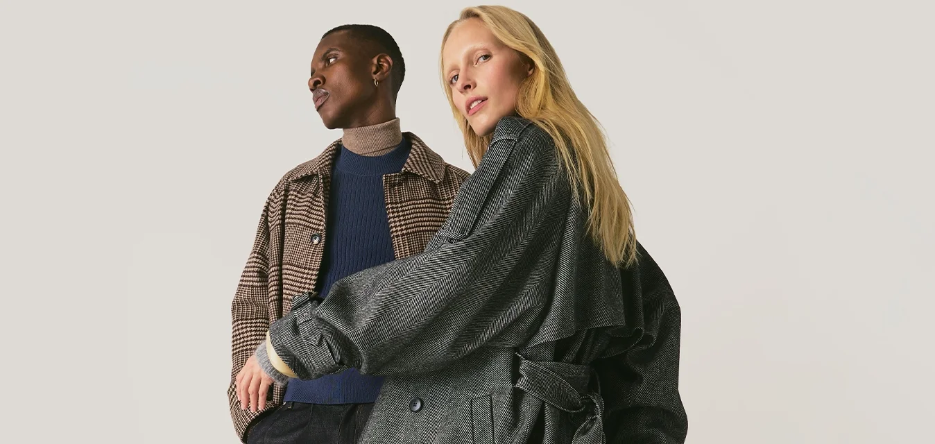 Due persone con capi esterni decorati sono in piedi contro uno sfondo chiaro uniforme, una con un cappotto lungo e fluente, l'altra con una giacca a quadri.