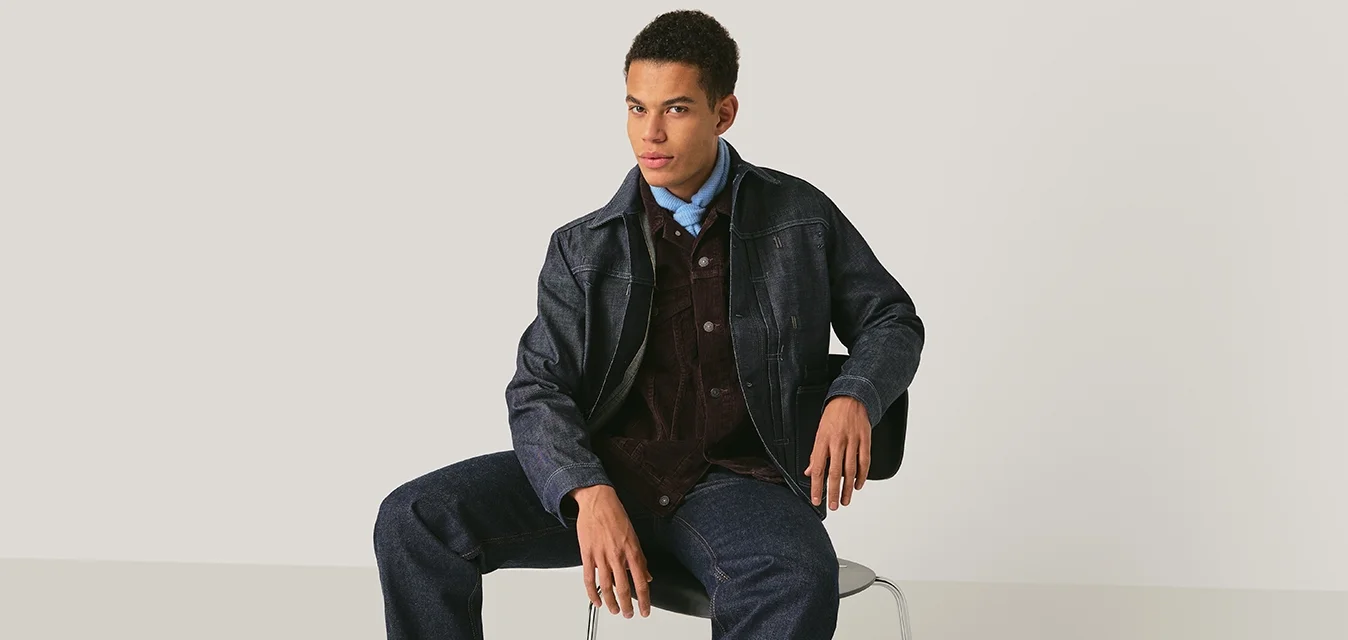 Giovane uomo con giacca scura a trama, camicia blu, gilet in velluto a coste, pantaloni di denim e stivali neri seduto su una sedia contro uno sfondo semplice.