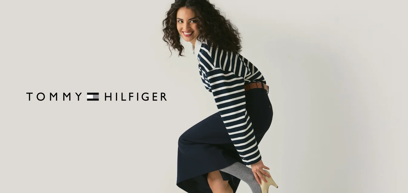 Donna sorridente, sistema un tacco alto beige sopra calzini grigi al ginocchio, indossa un maglione a righe e una gonna blu navy su uno sfondo chiaro e uniforme.