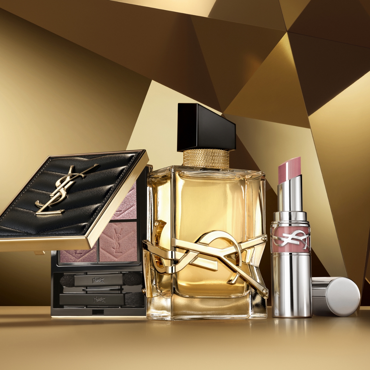 Un assortimento di prodotti di bellezza YSL, tra cui un compatto, un flacone di profumo e un rossetto, è elegantemente disposto su una superficie dorata riflettente con uno sfondo dorato geometrico.