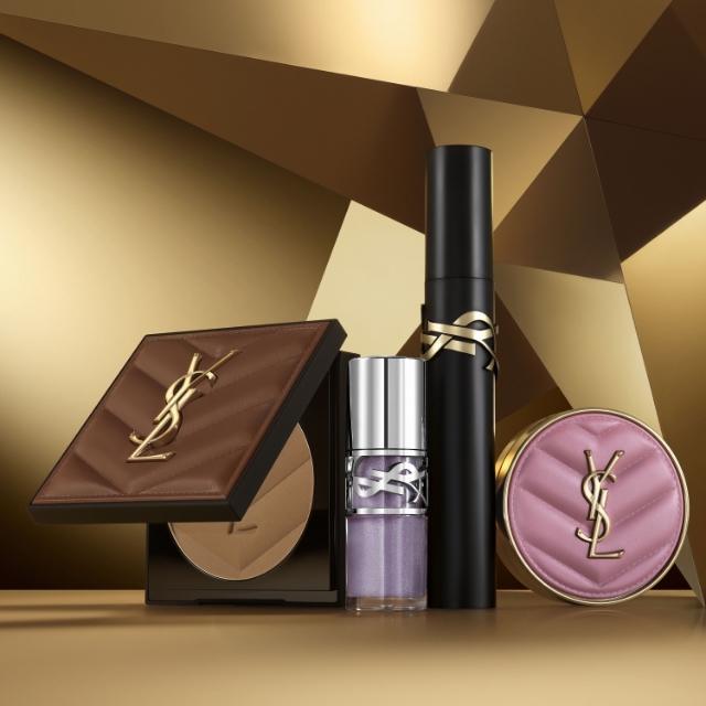 Un assortimento di prodotti di bellezza YSL, tra cui una palette compatta, un flacone di profumo e un rossetto, è disposto su una superficie dorata e geometrica.
