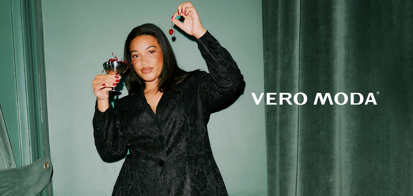 Blazer in pizzo nero e pantaloni svasati abbinati, con una texture liscia, tenuti da una persona con un bicchiere da cocktail e ciliegie. Tacchi rossi.