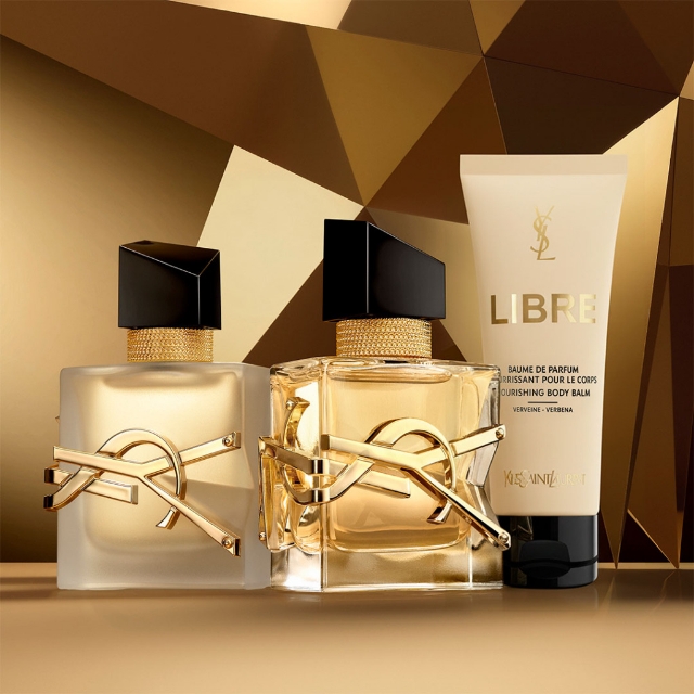 Due flaconi di profumo YSL Libre (uno satinato, uno trasparente) e un balsamo corpo nutriente Libre sono elegantemente esposti su uno sfondo dorato geometrico e dinamico.