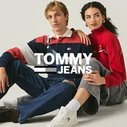 Un uomo con una maglietta a righe e salopette di denim è seduto accanto a una donna con un top rosso a maniche lunghe e pantaloni grigi su un pavimento bianco e semplice.