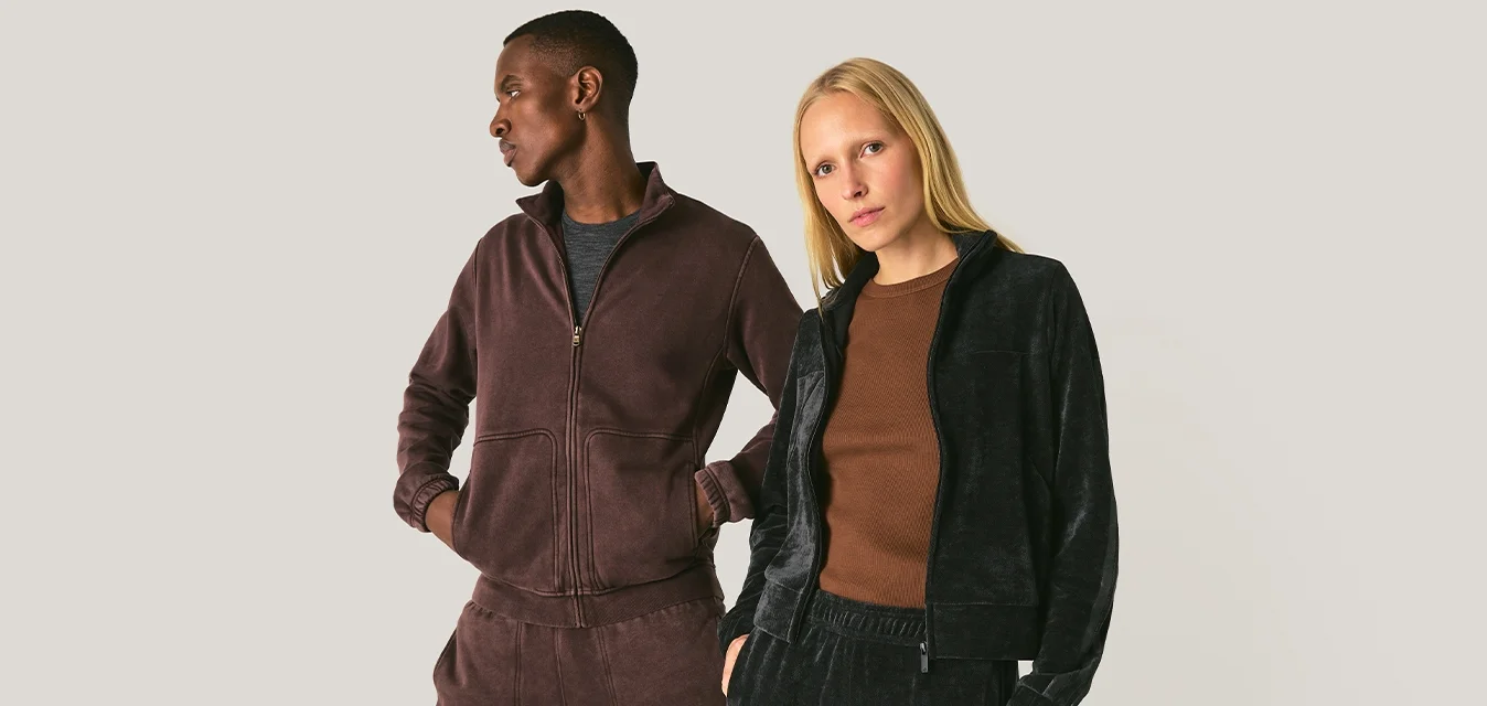 Due modelli in piedi fianco a fianco indossano tute casual in velluto, una marrone scuro e l'altra nera, su uno sfondo semplice.