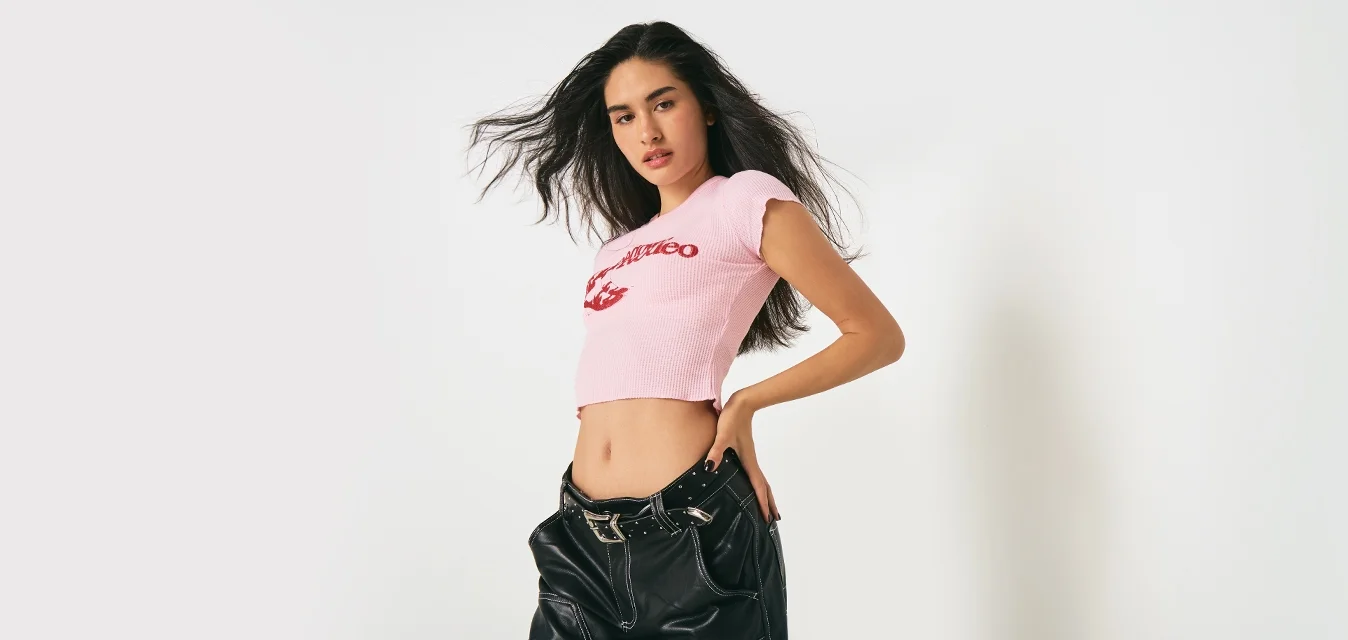 Giovane donna con capelli lunghi e scuri, indossa un top corto rosa e pantaloni di pelle nera, mentre posa su uno sfondo bianco semplice.