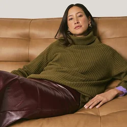 Maglione a collo alto verde oliva a coste, gonna in pelle bordeaux con una lucentezza, e un divano in pelle color cammello con schienale imbottito.
