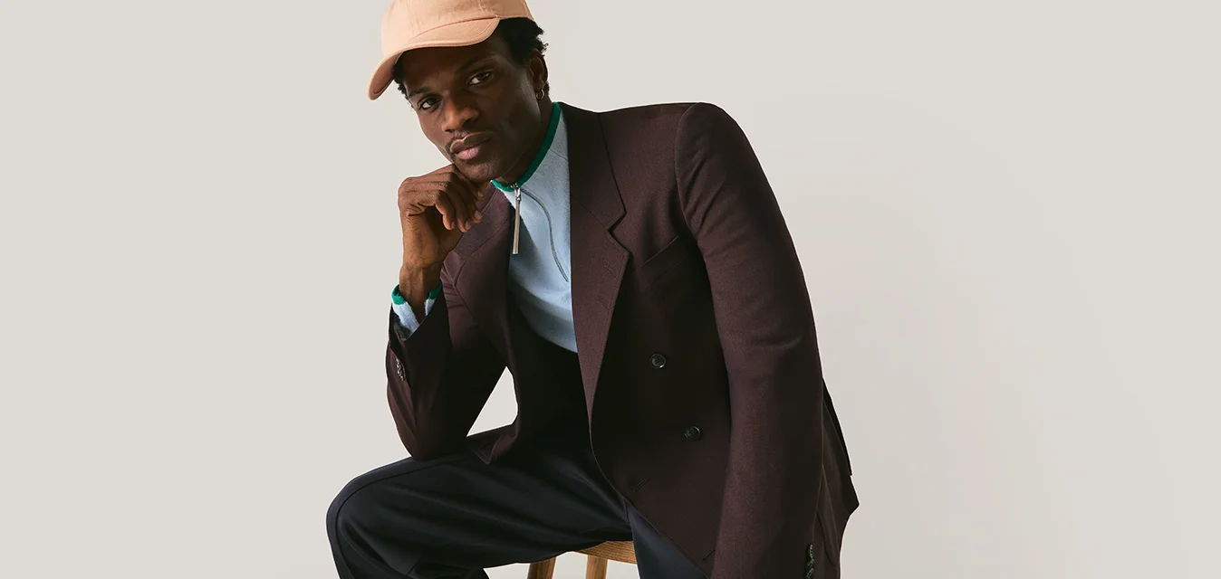 Uomo con cappello beige, blazer marrone, dolcevita blu, pantaloni neri e scarpe marroni seduto su uno sgabello di legno davanti a uno sfondo uniforme.