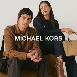 Uomo e donna seduti su un divano di tessuto color senape; l'uomo indossa una giacca marrone con una borsa nera a tracolla, la donna indossa un completo scuro con una borsa beige e stivali.