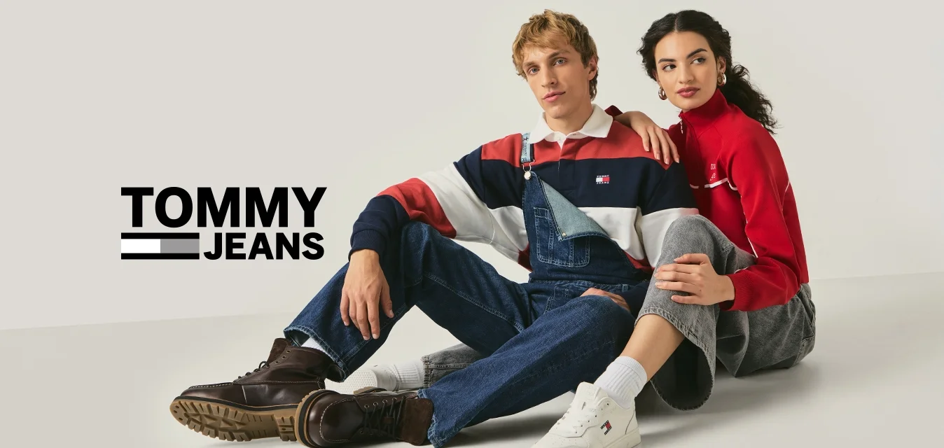 Un uomo con una maglietta a righe e salopette di denim è seduto accanto a una donna con un top rosso a maniche lunghe e pantaloni grigi su un pavimento bianco e semplice.