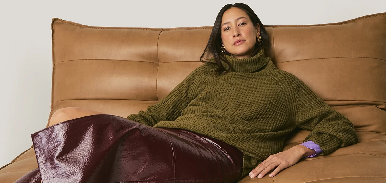 Maglione a collo alto verde oliva a coste, gonna in pelle bordeaux con una lucentezza, e un divano in pelle color cammello con schienale imbottito.