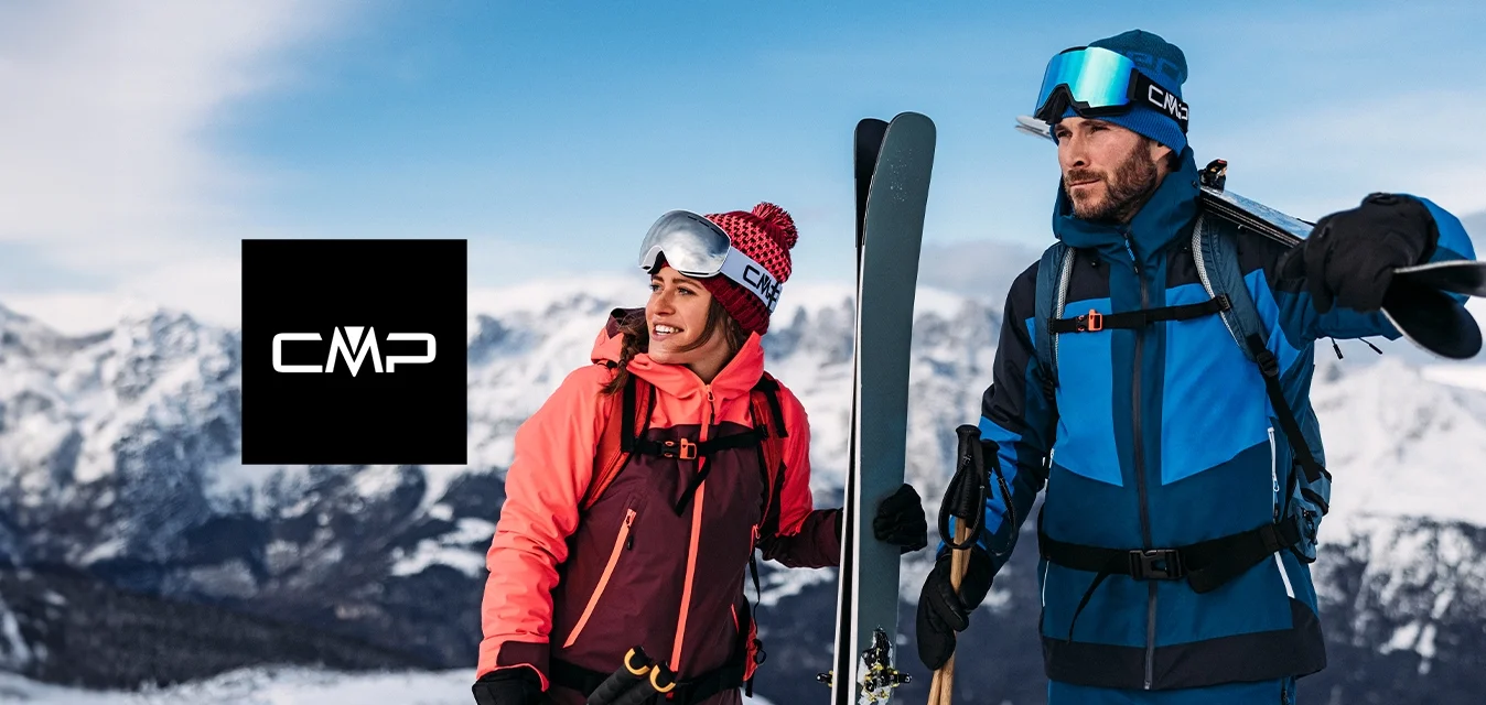 Due sciatori si trovano su un terreno innevato. La donna indossa una giacca rosa e marrone scuro, pantaloni rosa abbinati e un cappello di lana rosso. L'uomo è vestito con una giacca e pantaloni blu e neri, tiene in mano gli sci e i bastoncini. Entrambi indossano occhiali da sci.