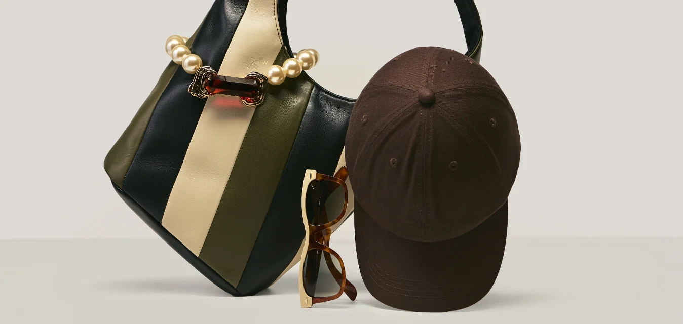 Borsa a righe con dettaglio di perle, cappellino da baseball marrone scuro e occhiali da sole tartarugati disposti su una superficie chiara e semplice.
