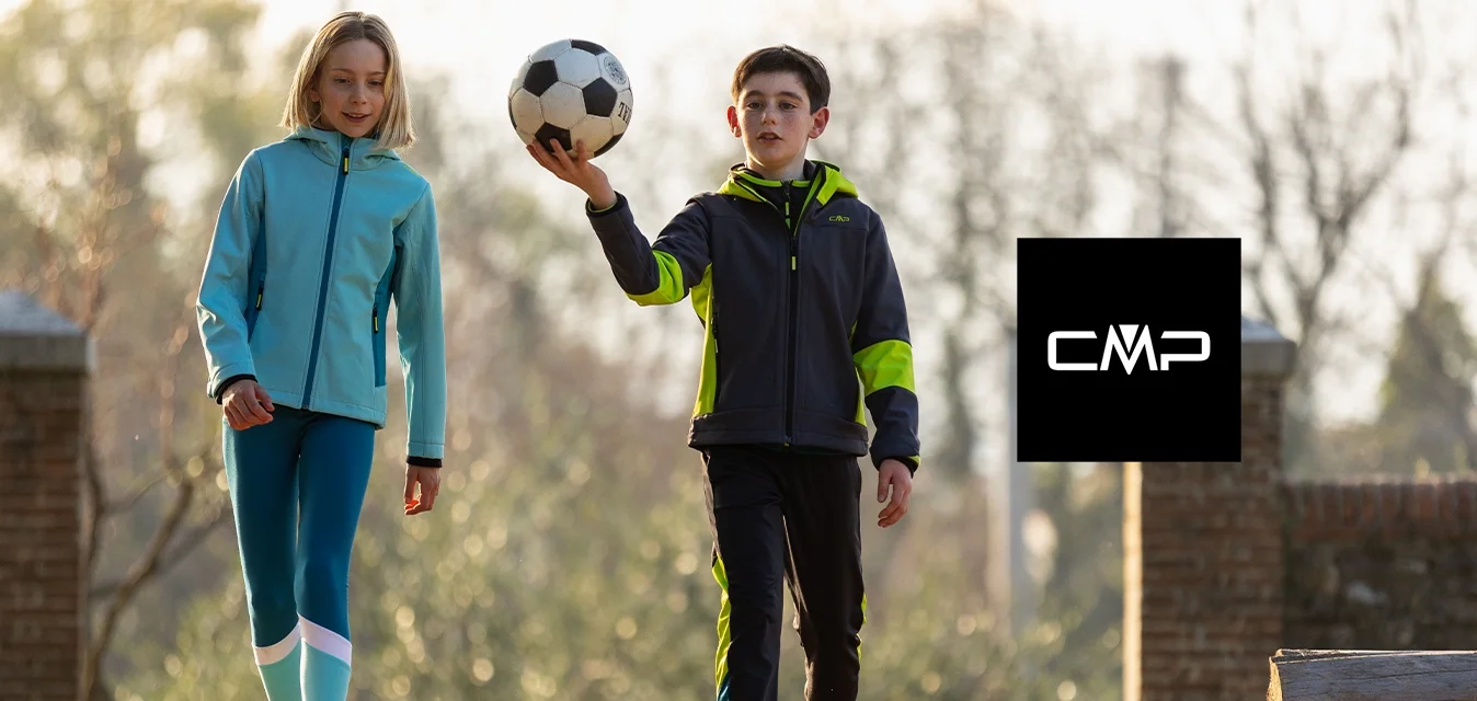 Due bambini camminano su un sentiero erboso, uno con una giacca teal e leggings blu, l'altro con una giacca grigia con accenti verdi, entrambi mentre tengono in mano un pallone da calcio.