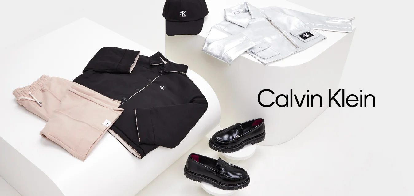 Giacca nera con bottoni, pantaloni jogger rosa pallido, giacca argentata con zip, cappellino nero e mocassini neri esposti su piattaforme geometriche bianche.