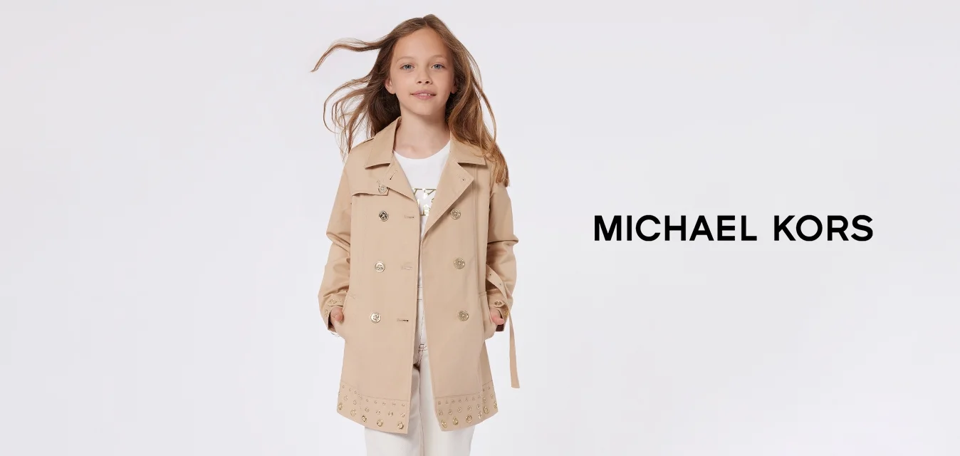 Trench coat beige doppiopetto con bottoni dorati e orlo borchiato, indossato su una maglietta bianca e pantaloni bianchi cropped, completato da sneakers bianche.