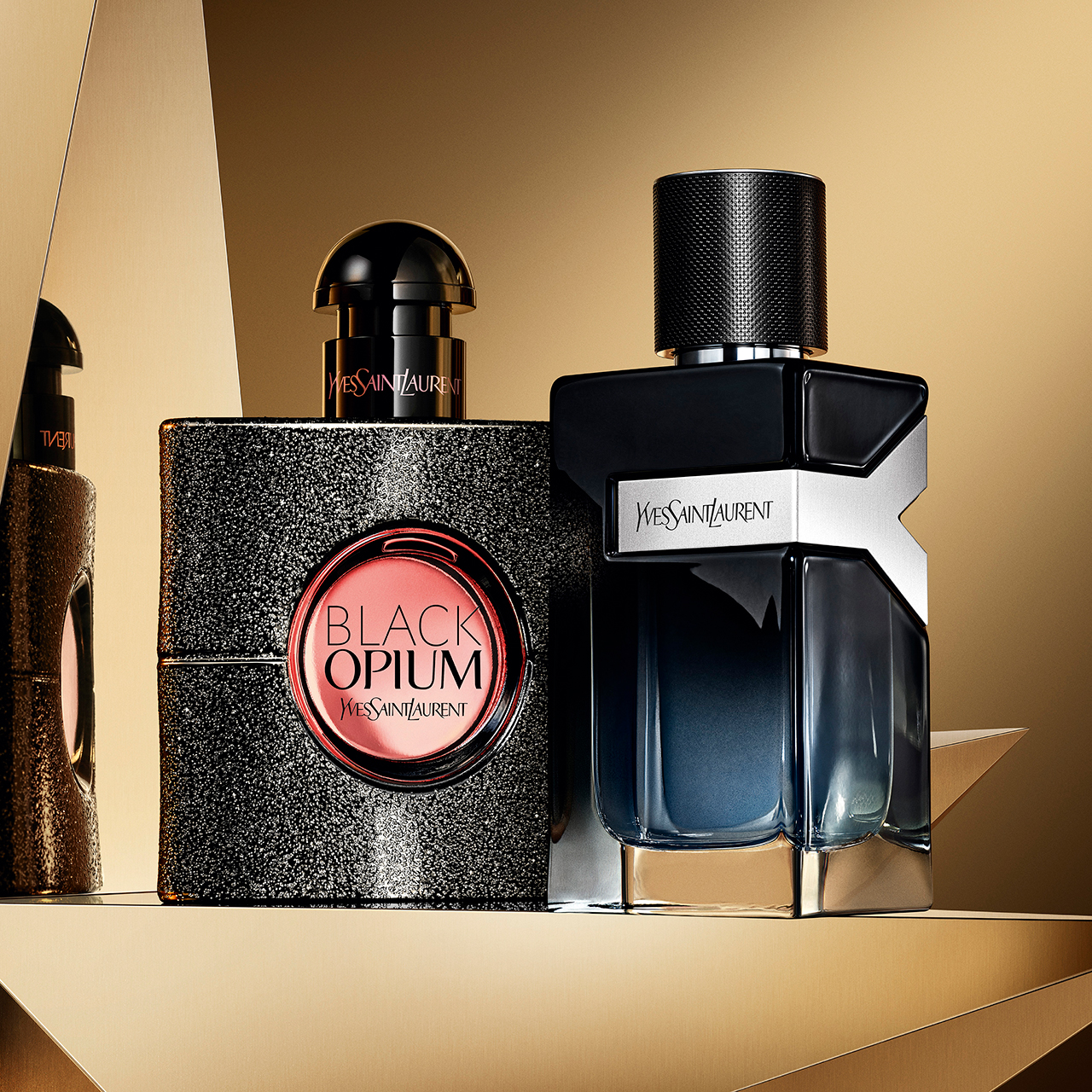 Due distinti profumi Yves Saint Laurent, una bottiglia alta nera di YSL Y Eau de Parfum e una bottiglia dorata di YSL Libre, sono elegantemente esposti su uno sfondo geometrico dorato dinamico.