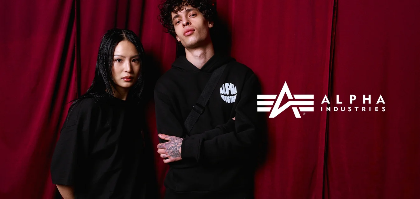 Due giovani adulti in abbigliamento casual nero stanno davanti a delle tende color bordeaux; uno di loro ha tatuaggi sulla mano e porta una borsa a tracolla.