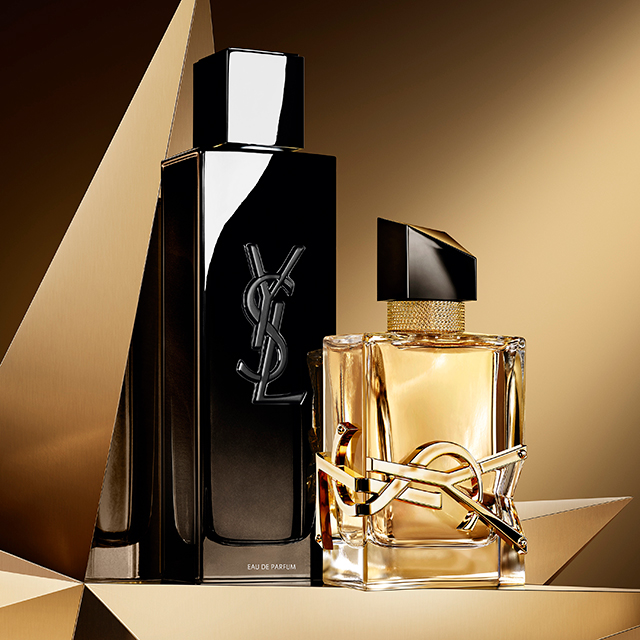 Tre eleganti confezioni regalo Yves Saint Laurent, due pi&ugrave; grandi e una pi&ugrave; piccola, con un logo YSL dorato e un motivo a strisce testurizzato, su uno sfondo geometrico dorato e scintillante.