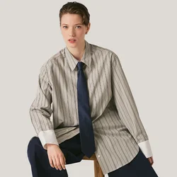 Giovane con capelli corti seduto su uno sgabello di legno, indossa una camicia a righe oversize, una cravatta blu navy, pantaloni larghi e scarpe nere.