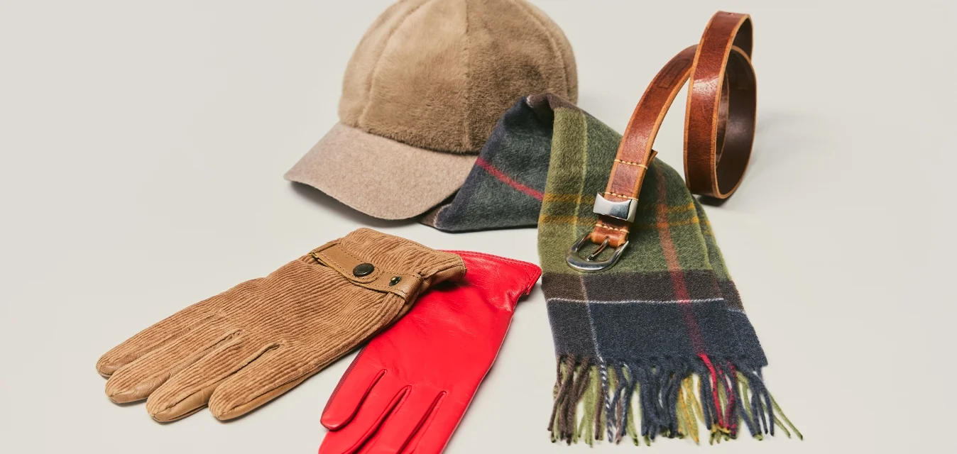 Berretto marrone, guanti di velluto a coste marroni, guanti in pelle rossa, sciarpa a quadri verde e blu, e cintura in pelle marrone su sfondo chiaro.