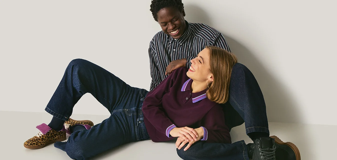 Due persone sedute su una superficie liscia. Una indossa un maglione viola scuro con colletto a contrasto, l'altra una camicia a righe. Diverse scarpe sono presenti nell'immagine.