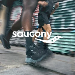 Scarpe sportive nere con una struttura testurizzata e suole grigie chiare in movimento su un pavimento, con uno sfondo urbano di graffiti sfocati.