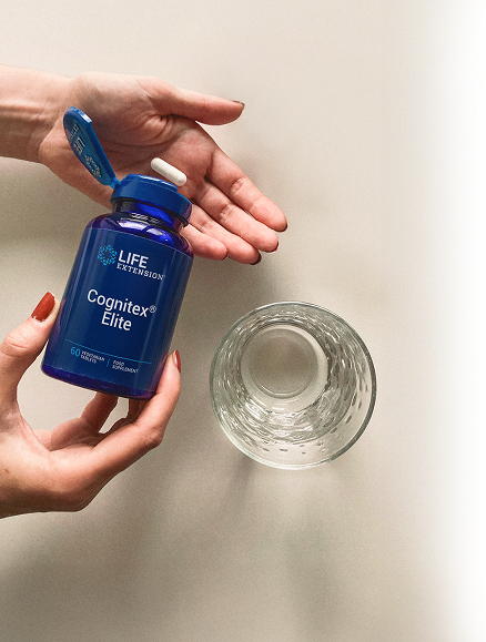 alt="Le mani di una donna che tengono una bottiglia di Life Extension Cognitex Elite in una mano e una capsula bianca nell'altra, accanto a un bicchiere d'acqua su una superficie di colore neutro.»