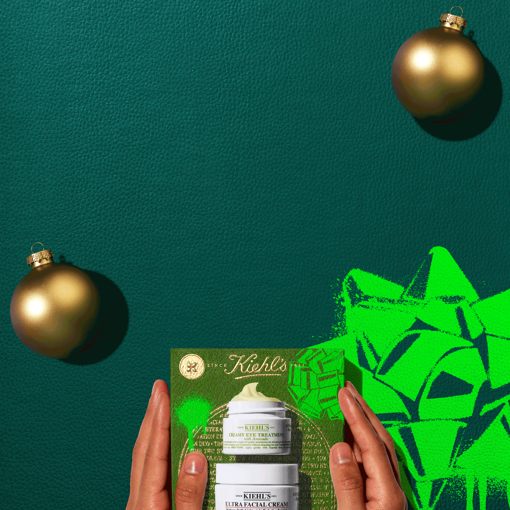 Confezione regalo Kiehl&rsquo;s verde con Creamy Eye Treatment e Ultra Facial Cream, tenuta da due mani su sfondo verde. In alto si vedono due palline natalizie dorate e in basso a destra un grande fiocco verde illustrato
