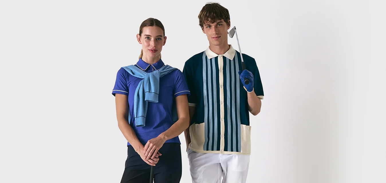 Due persone stanno fianco a fianco. La donna indossa una polo viola e pantaloni neri, mentre l'uomo ha una camicia a righe e pantaloni bianchi.