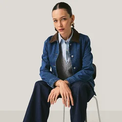 Giacca di jeans con colletto marrone, gilet grigio e camicia a righe azzurro chiaro. Jeans scuri a gamba larga e scarpe marroni con lacci. Seduto su una sedia.