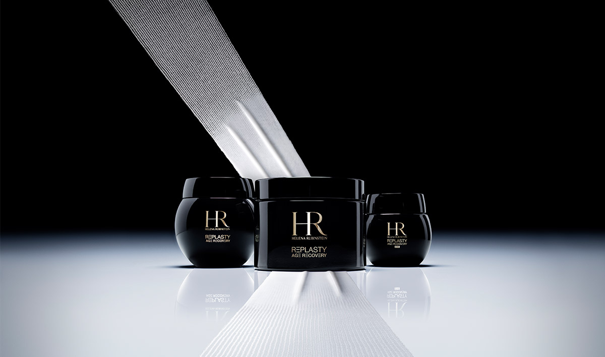 Presentazione della gamma completa Helena Rubinstein Replasty Age Recovery su una superficie riflettente. Si vedono tre vasi neri di diverse dimensioni allineati, accompagnati da una striscia bianca artistica che attraversa l'immagine in diagonale, ricordando il concetto di 'bendaggio' cosmetico.