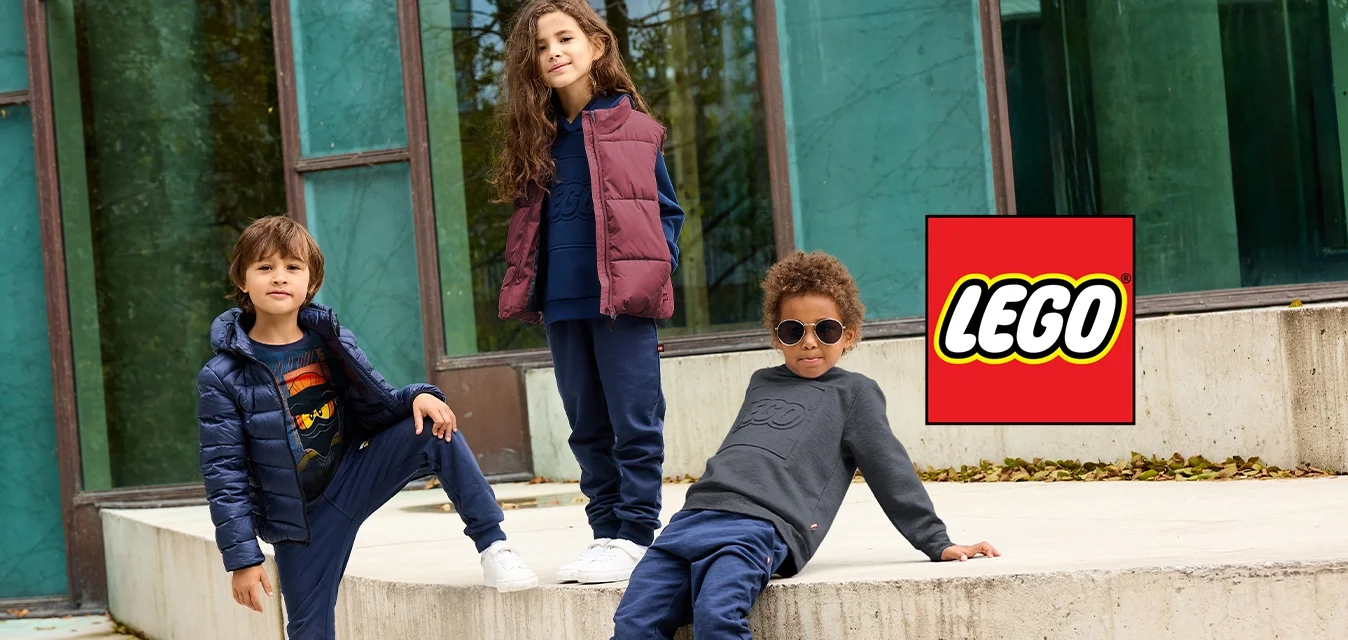 Tre bambini in abbigliamento autunnale informale posano all'aperto vicino a un moderno edificio in vetro, con un bambino che indossa occhiali da sole e si appoggia all'indietro.