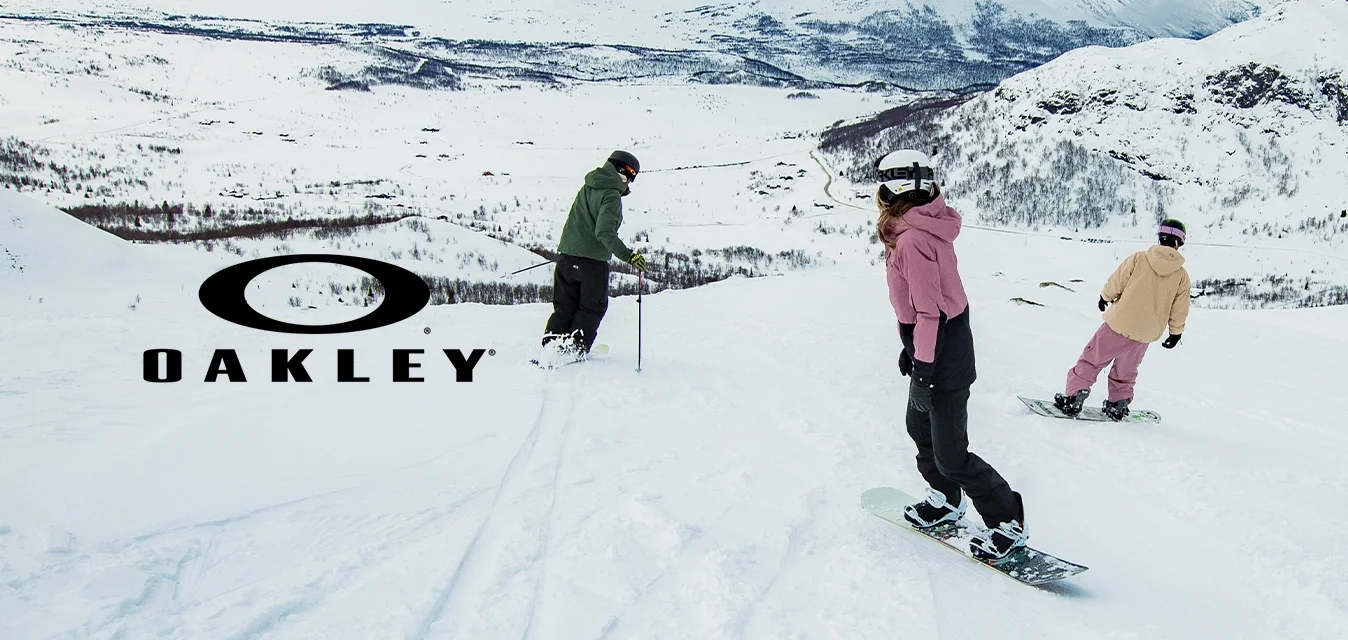 Tre snowboarder scendono lungo un pendio nevoso, indossando una giacca verde, un felpa rosa e una giacca beige. Lo sfondo presenta montagne coperte di neve.