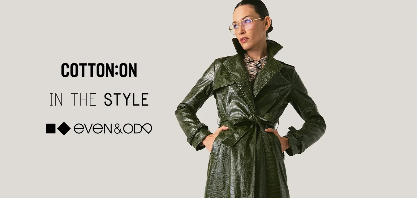 Donna con trench colore verde scuro con cintura, gonna marrone con spacco, stivali neri con punta e occhiali, in piedi con le mani sui fianchi.