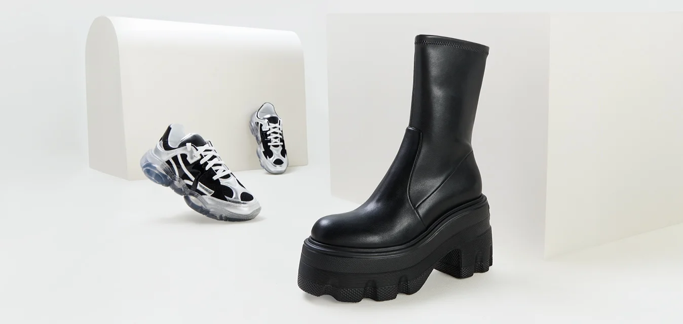 Stivali platform in pelle nera con suola spessa, texture liscia e design aderente, accanto a sneaker in bianco e nero su uno sfondo bianco.
