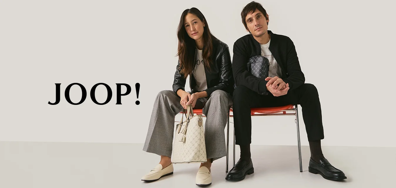 Due adulti seduti su sedie rosse contro uno sfondo neutro; la donna tiene una borsa beige, l'uomo una pochette nera con motivi, entrambi indossano abiti casual.