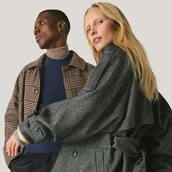 Due persone con capi esterni decorati sono in piedi contro uno sfondo chiaro uniforme, una con un cappotto lungo e fluente, l'altra con una giacca a quadri.