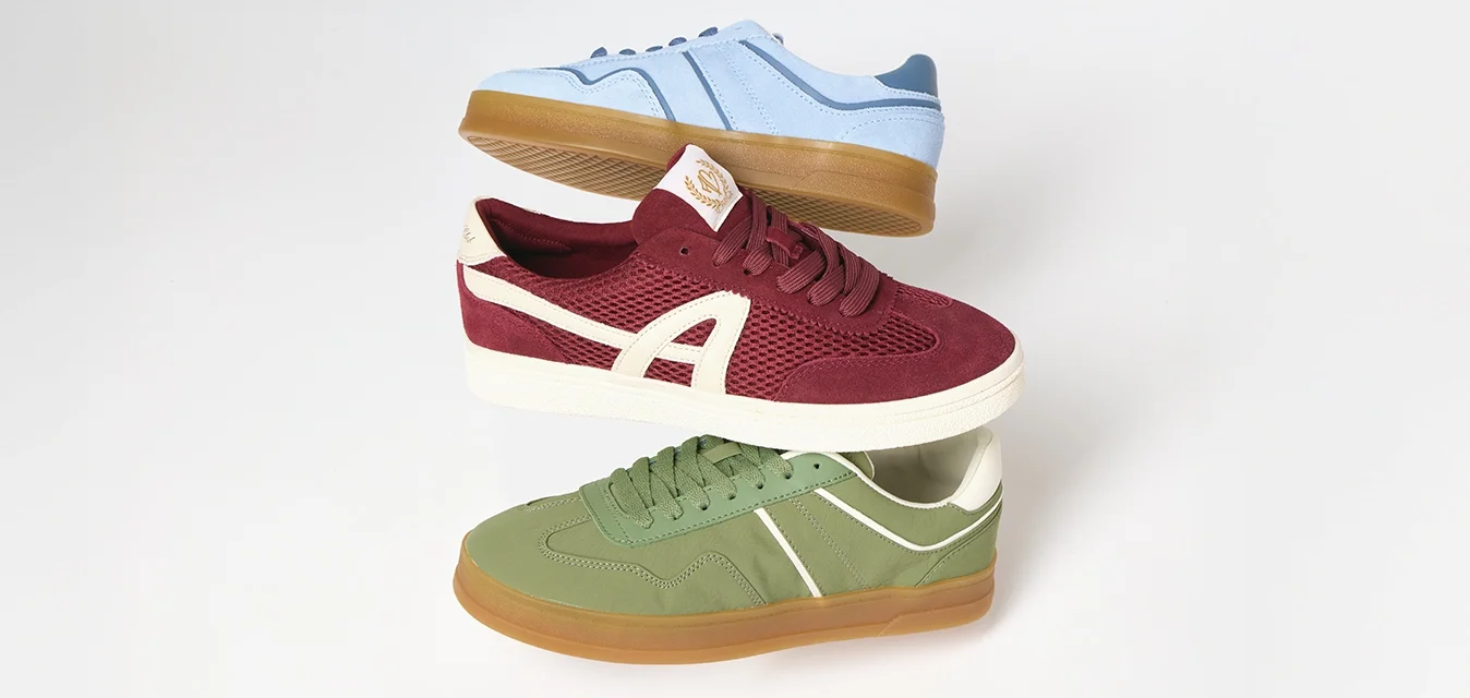 Tre scarpe da ginnastica impilate verticalmente: blu con suola in gomma, bordeaux con suola bianca e accenti, e verde con suola in gomma, su sfondo bianco.