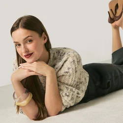 Donna sdraiata sul pavimento con il mento appoggiato sulle mani, indossa un maglione a motivo, pantaloni neri, tacchi marroni e braccialetti, su sfondo semplice.